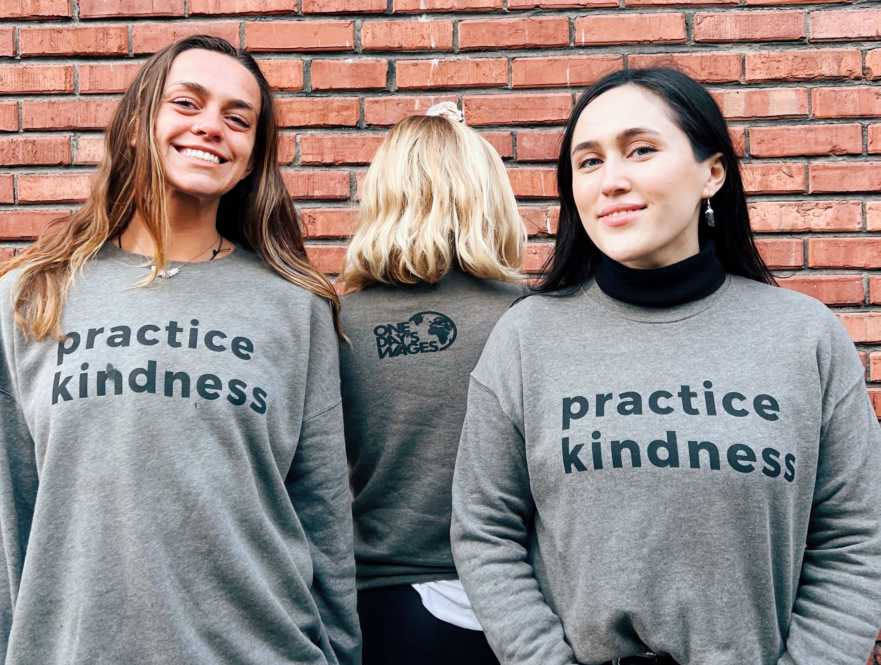 3 people stand with gray sweatshirts on that say Practice Kindness in dark gray bold font. One has the One Day's Wages logo on the back